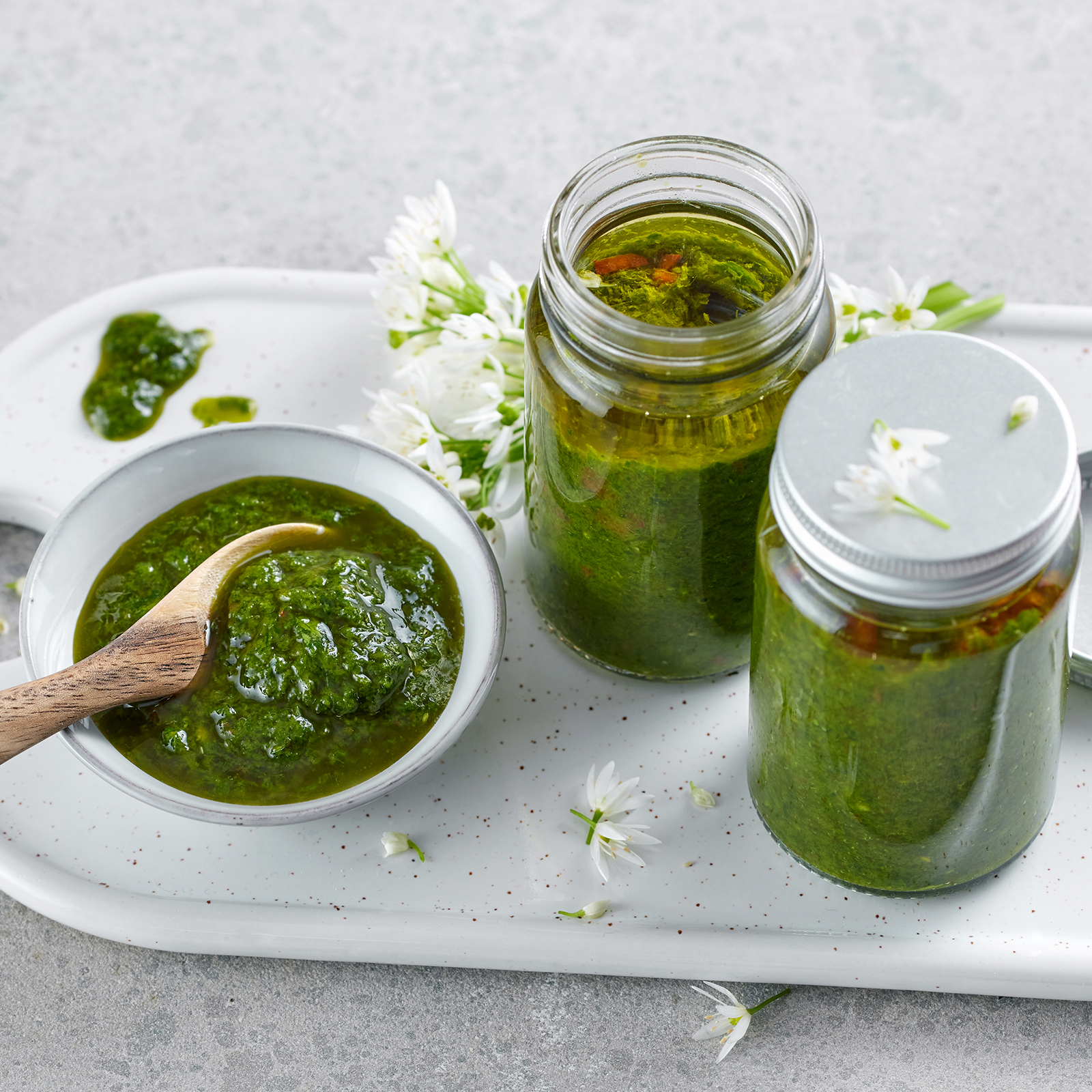 Bärlauch-Basilikum-Paste in einer Schale und zwei Einmachgläsern, dekoriert mit Bärlauchblüten auf einem weißen Porzellantablett.