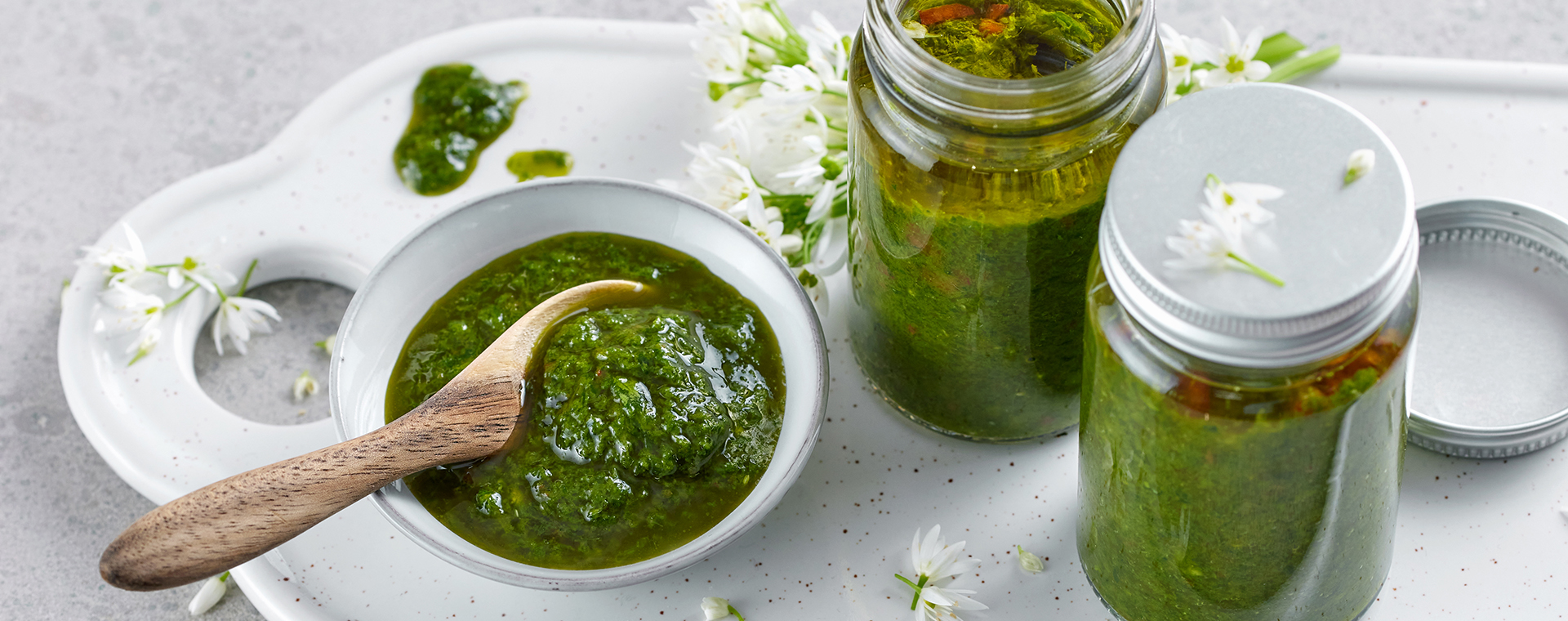 Bärlauch-Basilikum-Paste in einer Schale und zwei Einmachgläsern, dekoriert mit Bärlauchblüten auf einem weißen Porzellantablett.