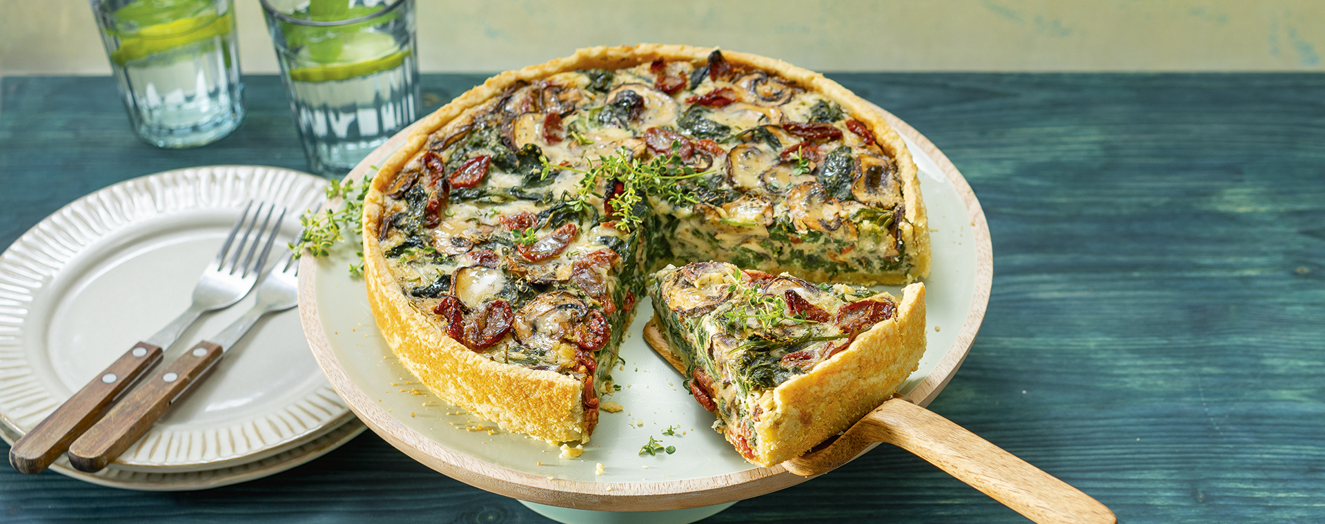 Champignon-Spinat-Quiche mit weißen Bohnen und getrockneten Tomaten auf einer Tortenplatte.