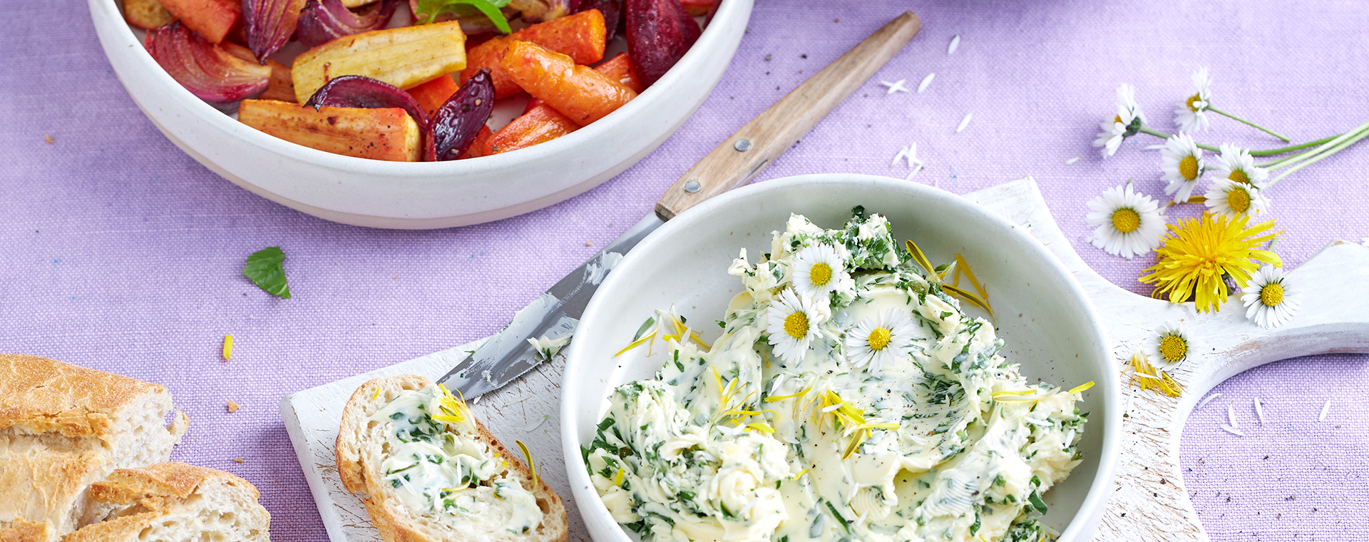 Wildkräuterbutter mit Gänseblümchen in einer Schale, dazu Baguette und Ofengemüse.